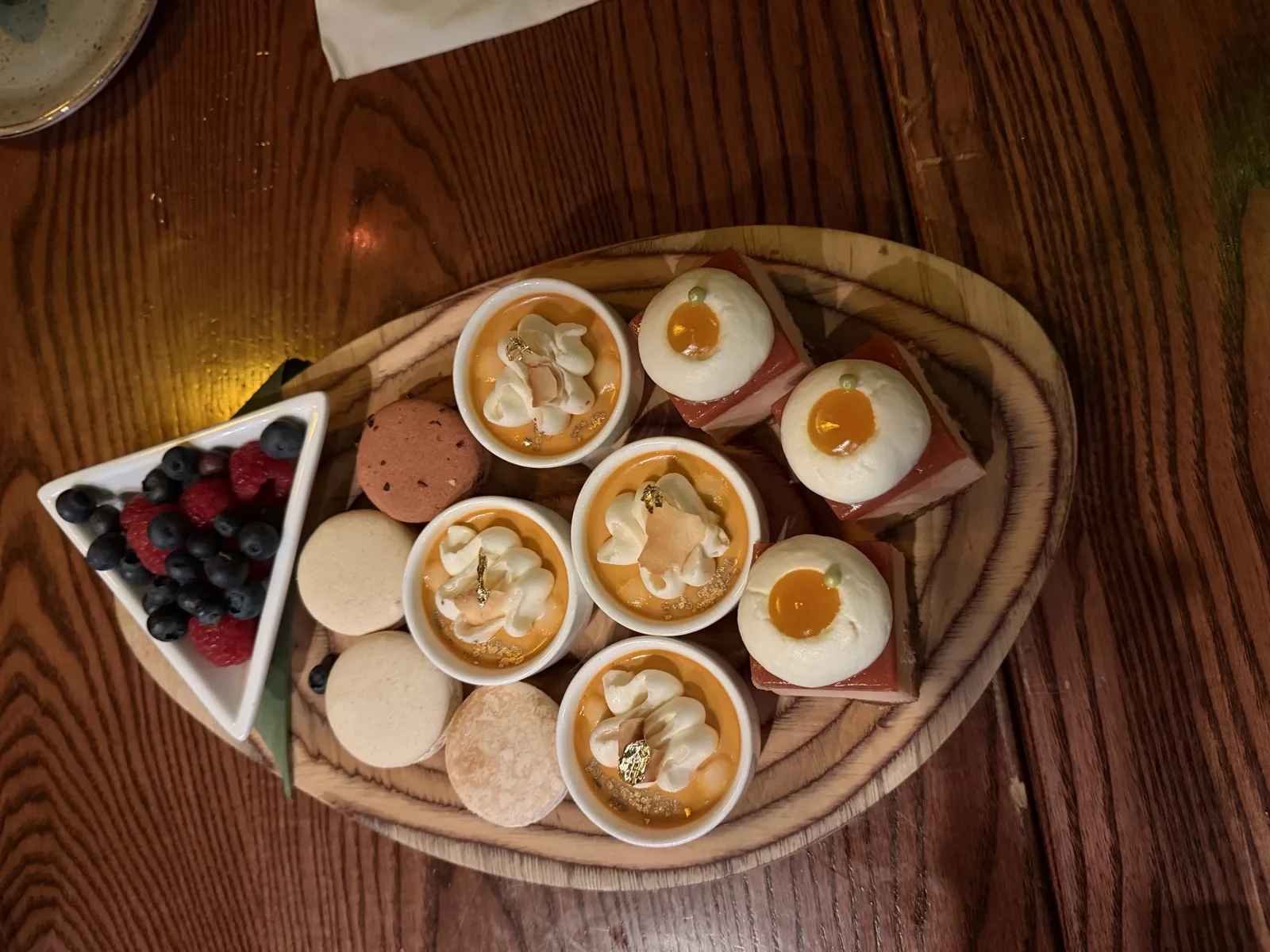 Overhead view of a wooden dessert board at the Tonga Room with macarons, salted caramel pots topped with whipped cream and gold leaf, mini cakes, and a bowl of fresh berries